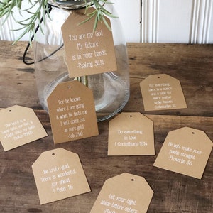 Pu&ograve; includere: Un set di otto etichette di carta marrone con testo bianco, ciascuna con una citazione ispiratrice diversa dalla Bibbia. Le etichette sono legate con uno spago e pendono da un barattolo di vetro pieno di verde.