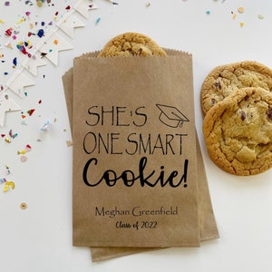 May include: A brown paper bag with the text "She's One Smart Cookie!" and the name "Meghan Greenfield" and "Class of 2022". Two chocolate chip cookies are on the table next to the bag.
