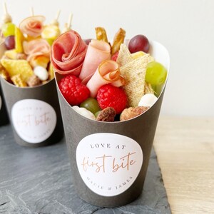 Op de afbeelding: Een zwarte papieren kegel gevuld met een verscheidenheid aan snacks, waaronder fruit, kaas, crackers en vlees. De kegel heeft een witte sticker met de tekst "LOVE AT first bite MACIE & JAMES".