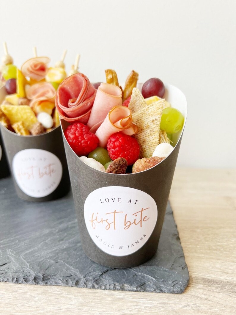 Op de afbeelding: Een zwarte papieren kegel gevuld met een verscheidenheid aan snacks, waaronder fruit, kaas, crackers en vlees. De kegel heeft een witte sticker met de tekst "LOVE AT first bite MACIE & JAMES".