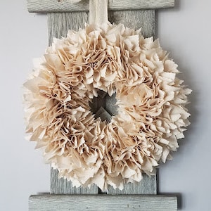 May include: A beige paper flower wreath hangs on a rustic wooden wall decor with a white ribbon bow.