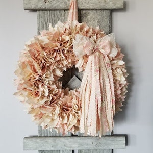 Puede incluir: Una corona de tela rosa y blanca con un lazo de tela rosa y blanca colgando de una estanter&iacute;a de pared de madera gris.