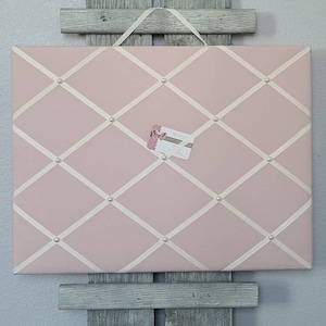May include: A pink fabric bulletin board with white ribbon crisscrossing the surface. The board is decorated with white pearl-like buttons and has a small card attached to it.