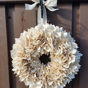 May include: A cream-colored fabric wreath with a ruffled texture, hanging from a brown wooden door with a white ribbon.