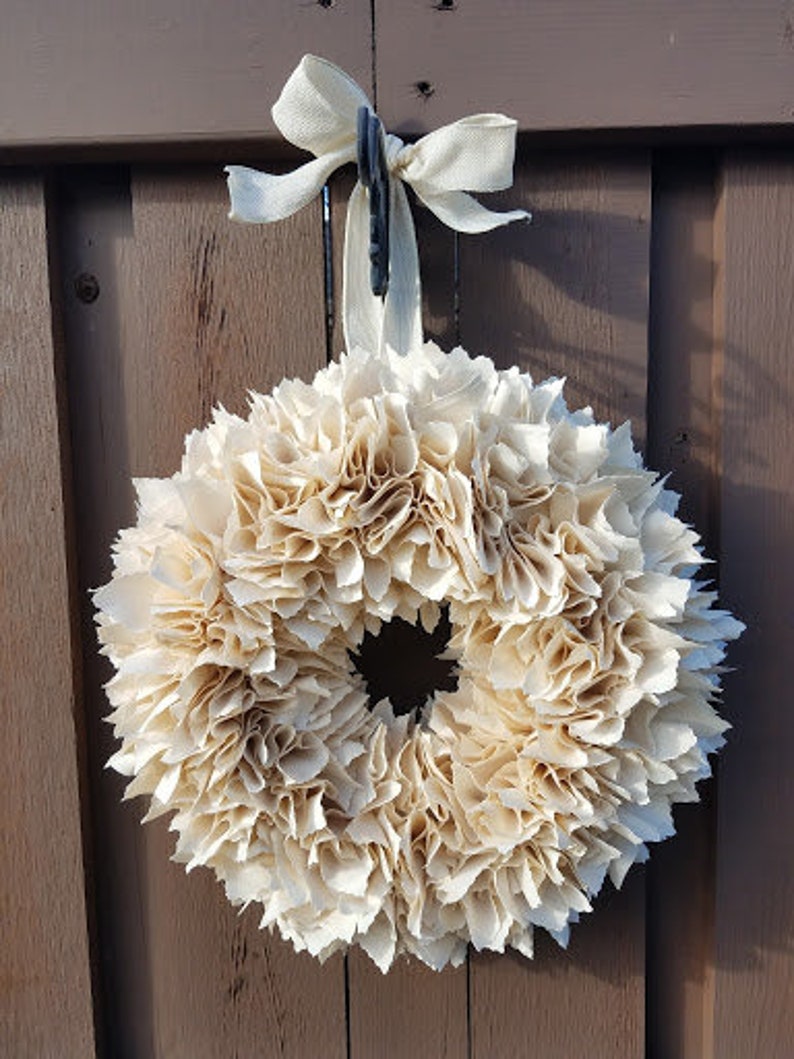 May include: A cream-colored fabric wreath with a ruffled texture, hanging from a brown wooden door with a white ribbon.