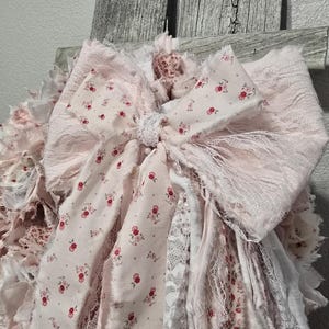 May include: A pink and white fabric garland with a large bow. The garland is made of various textures, including lace and floral print fabric. The bow is tied with a white ribbon.