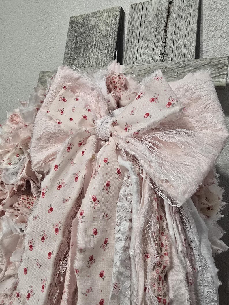 May include: A pink and white fabric garland with a large bow. The garland is made of various textures, including lace and floral print fabric. The bow is tied with a white ribbon.