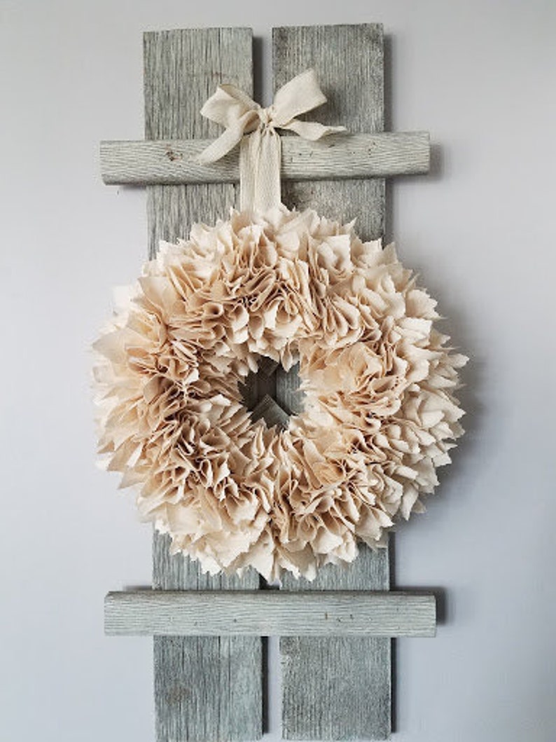 May include: A beige paper flower wreath hangs on a rustic wooden wall decor with a white ribbon bow.