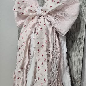 May include: A pink and white fabric bow with a floral pattern and lace trim. The bow is attached to a string of fabric strips with frayed edges.