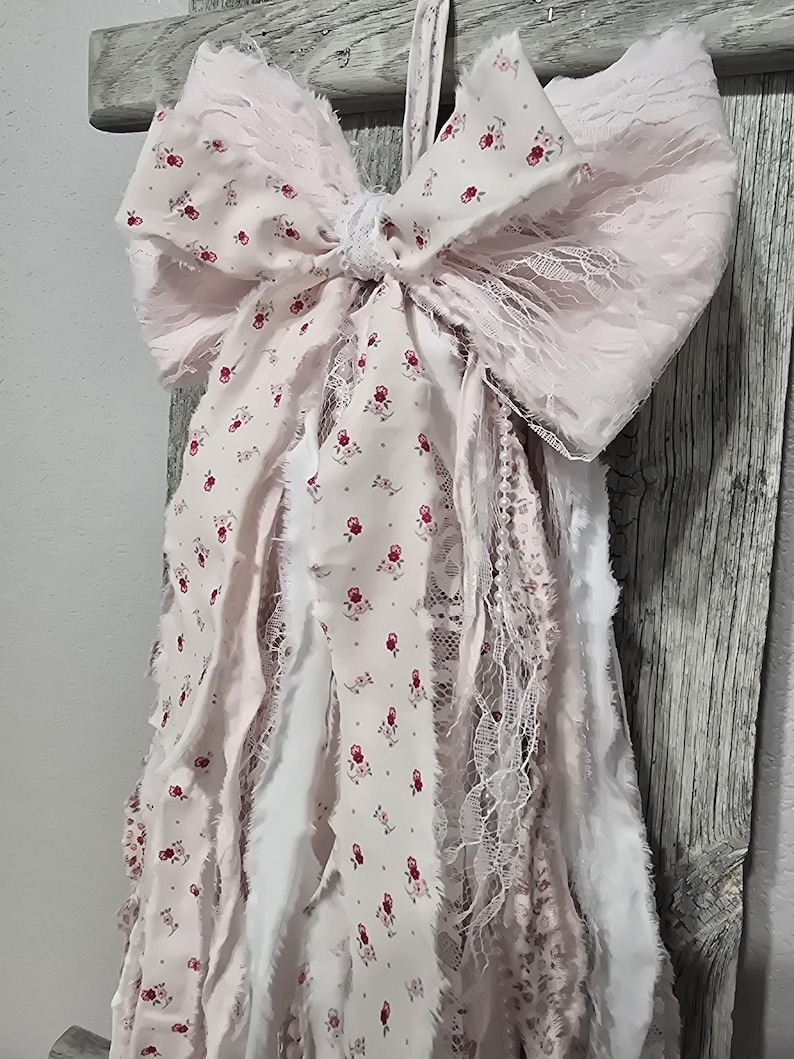 May include: A pink and white fabric bow with a floral pattern and lace trim. The bow is attached to a string of fabric strips with frayed edges.