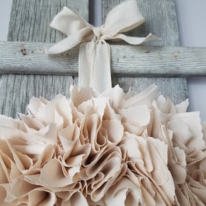 May include: A white, rustic wooden cross with a cream-colored fabric flower attached to the center. The flower is made of many layers of fabric, creating a fluffy, ruffled look. A white ribbon is tied around the cross, adding a touch of elegance.