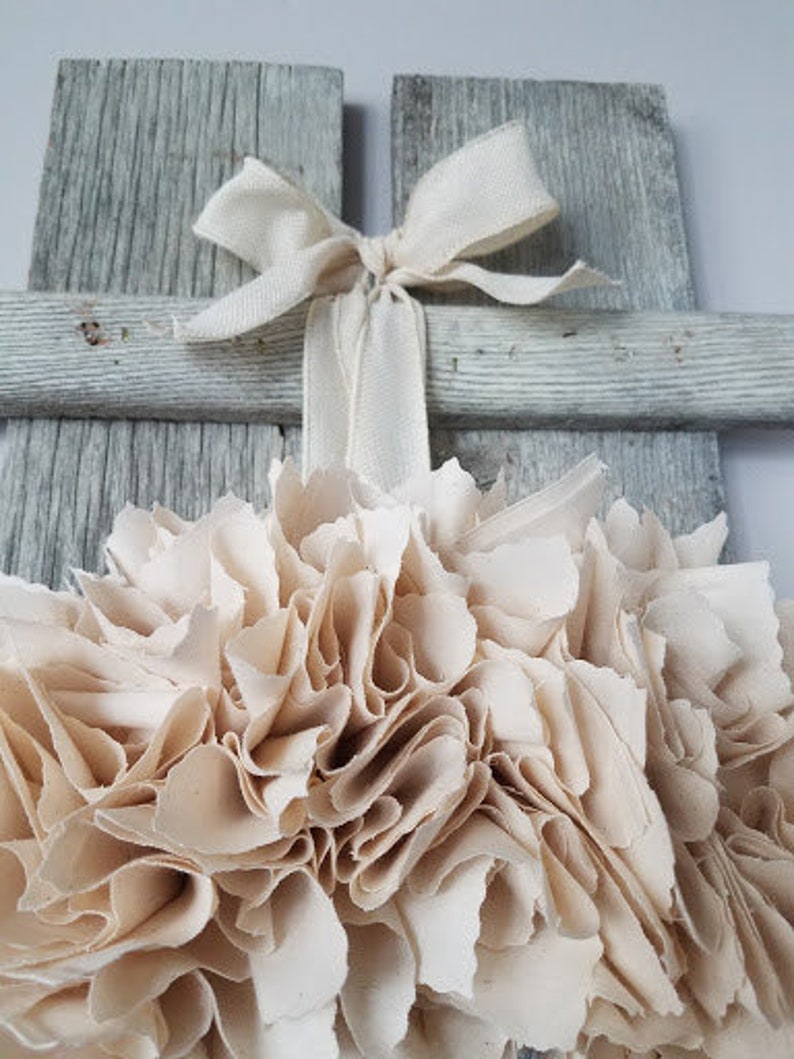 May include: A white, rustic wooden cross with a cream-colored fabric flower attached to the center. The flower is made of many layers of fabric, creating a fluffy, ruffled look. A white ribbon is tied around the cross, adding a touch of elegance.