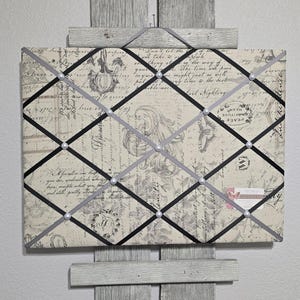 May include: A beige and black fabric bulletin board with a grid pattern of black and gray ribbon. The board is covered in a vintage-style print with cursive writing and floral designs. The ribbon is tied in a diamond pattern and secured with pearl-like buttons.