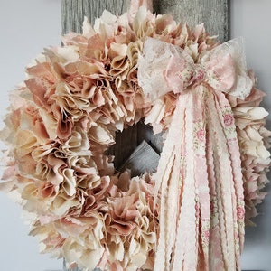 Puede incluir: Una corona de tela rosa y crema con un gran lazo de encaje rosa y blanco. La corona est&aacute; hecha de recortes de tela y es perfecta para una decoraci&oacute;n de estilo farmhouse o shabby chic.