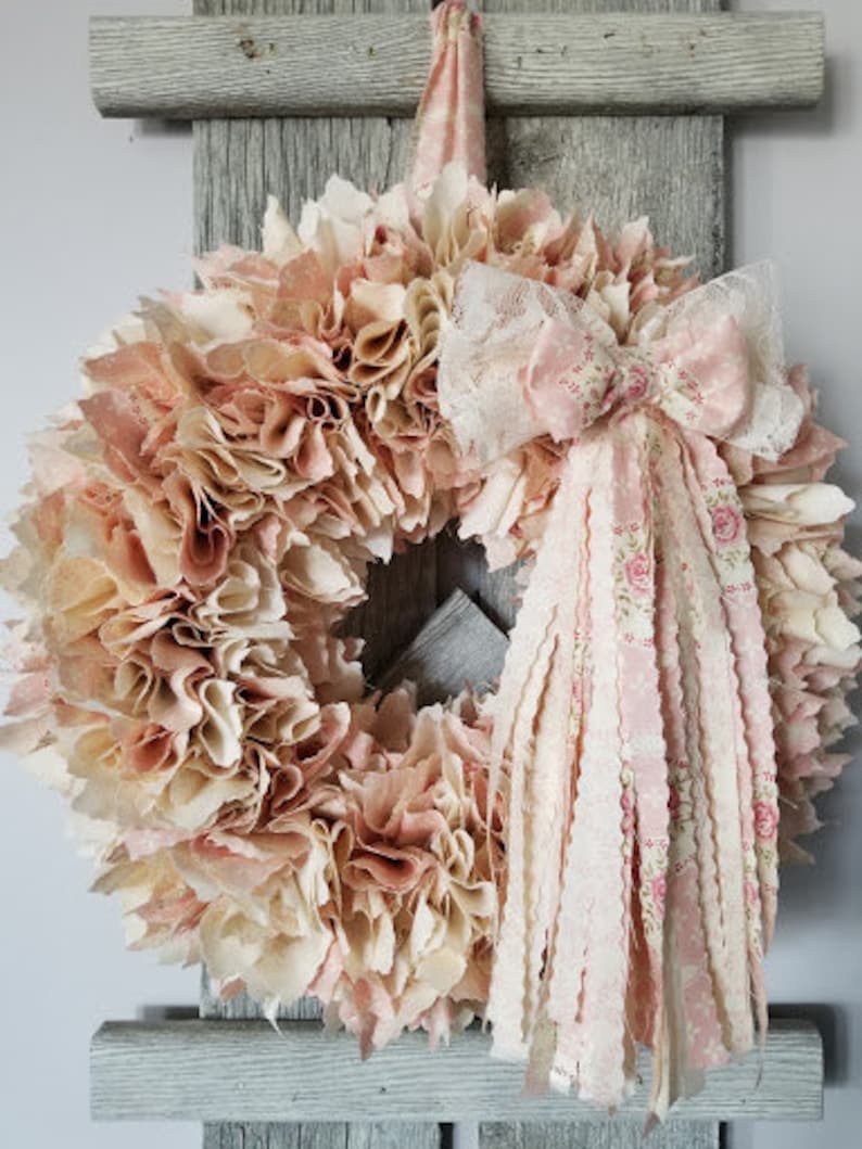 Puede incluir: Una corona de tela rosa y crema con un gran lazo de encaje rosa y blanco. La corona est&aacute; hecha de recortes de tela y es perfecta para una decoraci&oacute;n de estilo farmhouse o shabby chic.