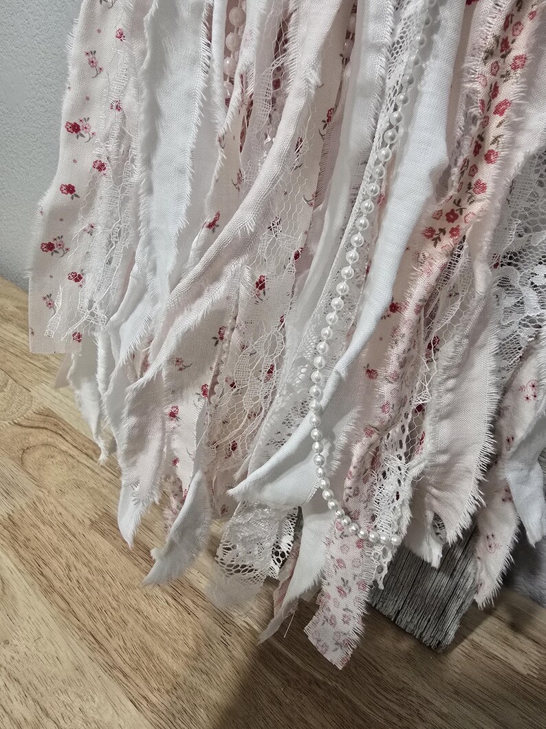 May include: A white and pink fabric garland with a pearl strand. The fabric is cut into strips and has a floral pattern.