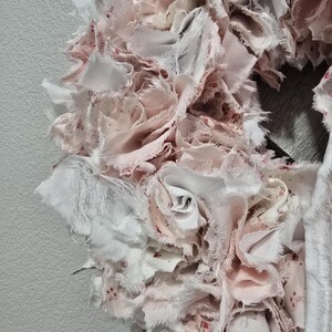 May include: A pink and white fabric wreath with a ruffled texture. The wreath is made of many small pieces of fabric that have been sewn together to create a layered effect.