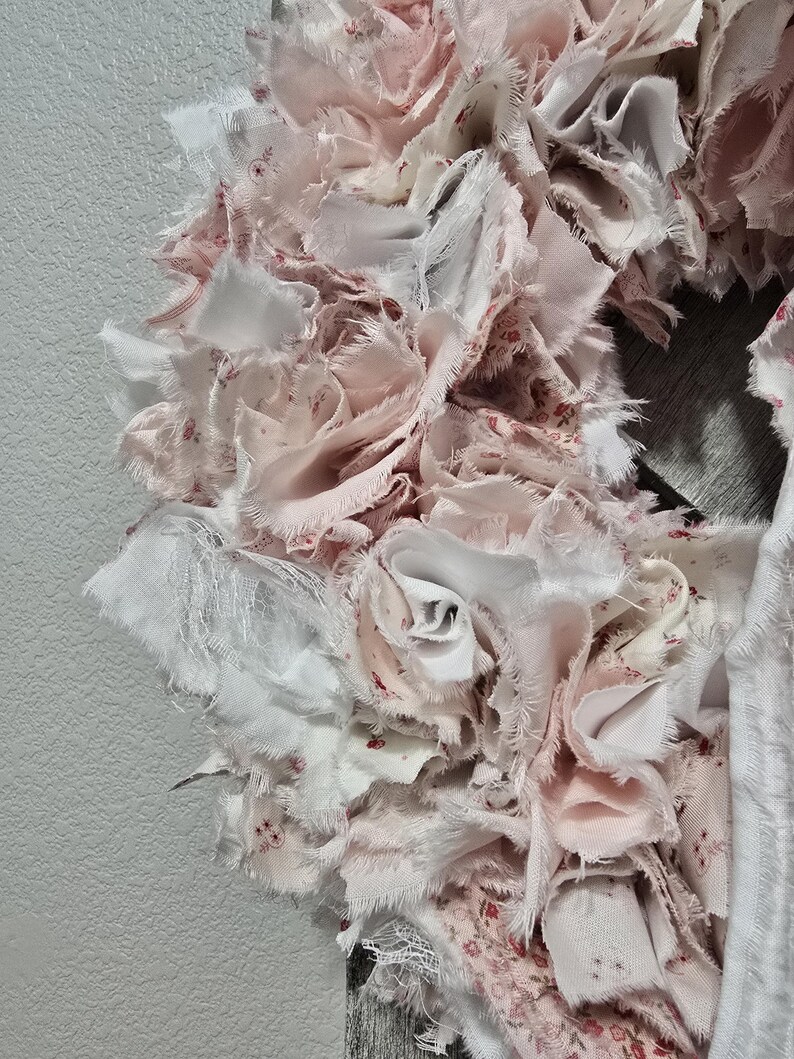 May include: A pink and white fabric wreath with a ruffled texture. The wreath is made of many small pieces of fabric that have been sewn together to create a layered effect.