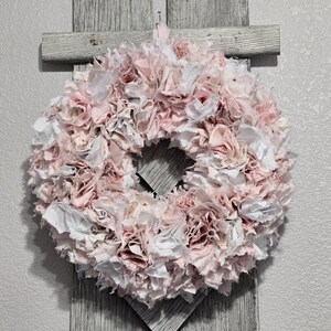 May include: A pink and white fabric wreath with a ruffled texture. The wreath is hanging on a gray wooden background.