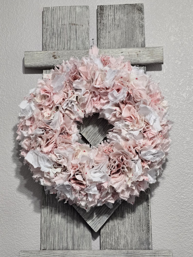 May include: A pink and white fabric wreath with a ruffled texture. The wreath is hanging on a gray wooden background.