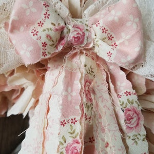 Puede incluir: Una corona de tela rosa y blanca con un gran lazo rosa y estampado floral. La corona est&aacute; hecha de tiras de tela con bordes deshilachados.