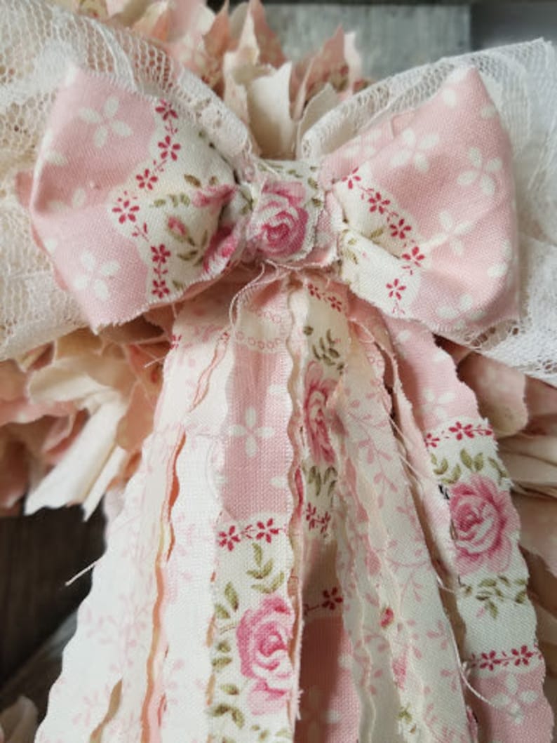 Puede incluir: Una corona de tela rosa y blanca con un gran lazo rosa y estampado floral. La corona est&aacute; hecha de tiras de tela con bordes deshilachados.