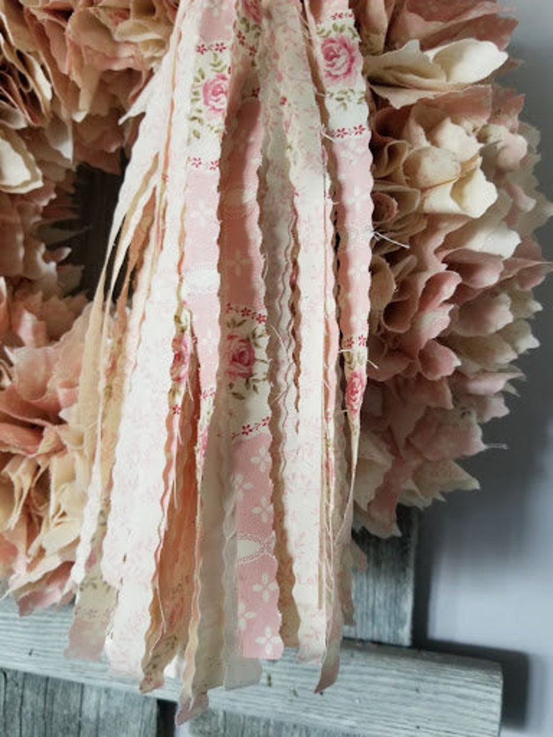 Puede incluir: Una corona de tejido rosa y blanco con una larga borla de tejido deshilachado que cuelga de la parte inferior. La borla est&aacute; hecha de m&uacute;ltiples tiras de tejido con un patr&oacute;n floral.