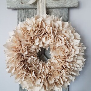 May include: A beige fabric wreath with a ruffled texture. The wreath is hanging on a rustic wooden ladder.