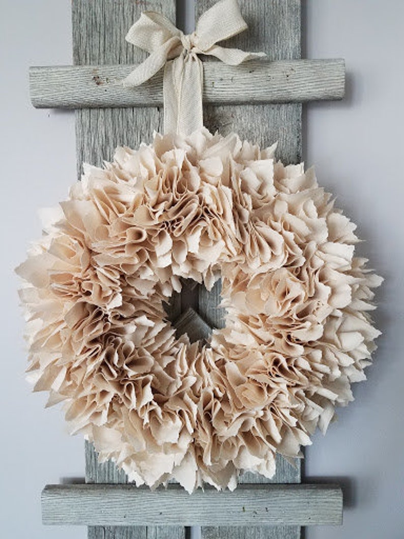 May include: A beige fabric wreath with a ruffled texture. The wreath is hanging on a rustic wooden ladder.