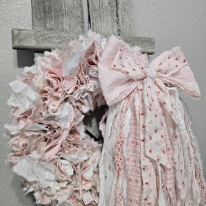 May include: A pink and white fabric wreath with a large pink bow. The wreath is made of strips of fabric that have been frayed and tied together. The bow is made of the same fabric and has a white center.
