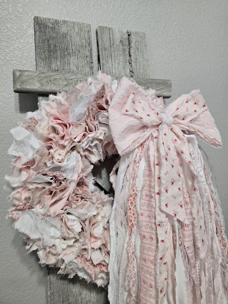 May include: A pink and white fabric wreath with a large pink bow. The wreath is made of strips of fabric that have been frayed and tied together. The bow is made of the same fabric and has a white center.