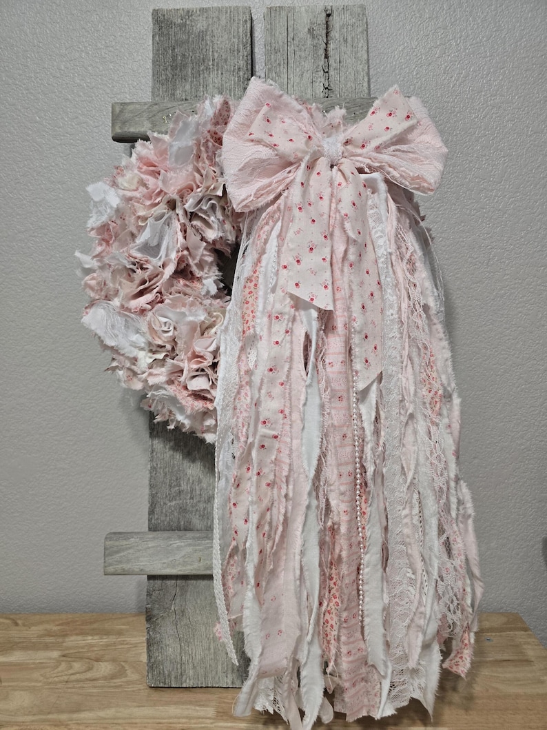 May include: A pink and white fabric wreath with a large pink bow. The wreath is made of strips of fabric and lace, and the bow is made of a patterned fabric. The wreath is hanging on a wooden board.