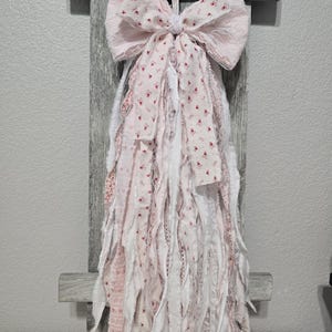 May include: A pink and white fabric bow with frayed edges and a floral pattern. The bow is hanging from a wooden surface.