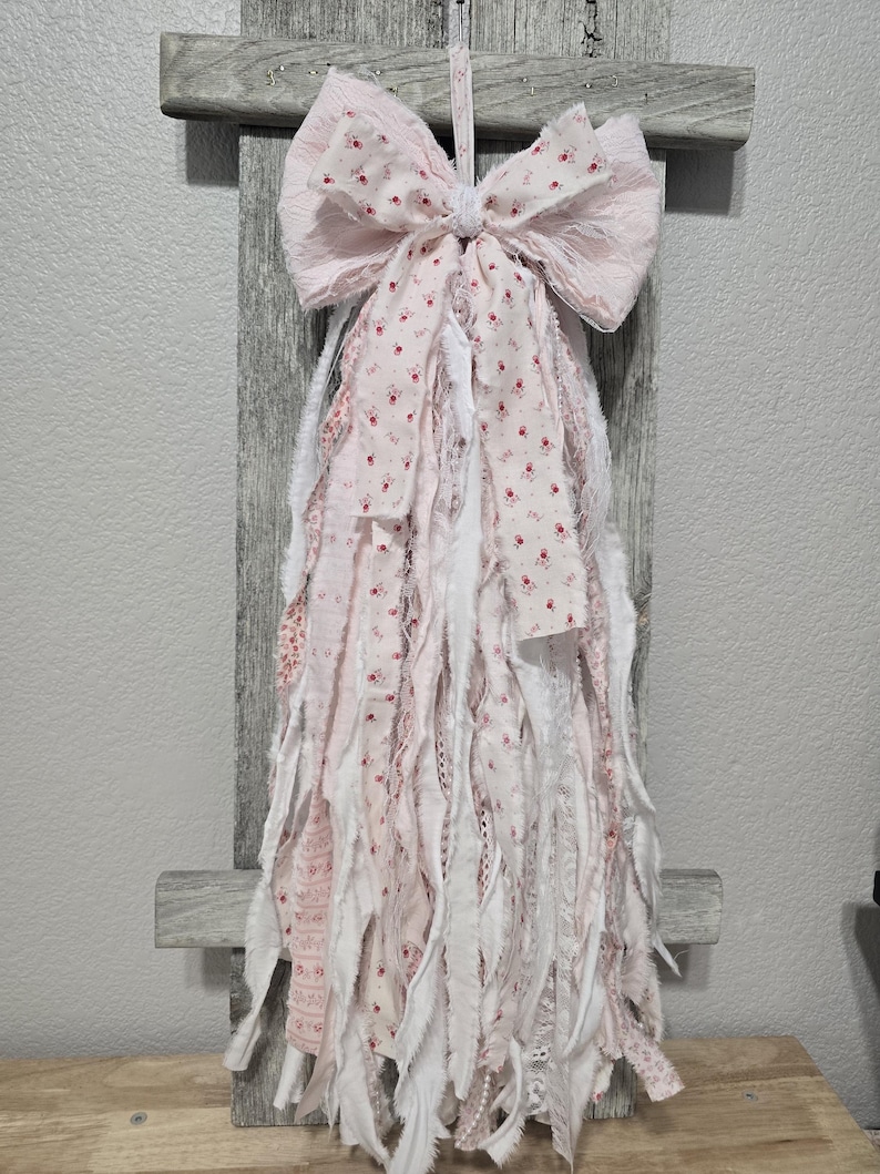 May include: A pink and white fabric bow with frayed edges and a floral pattern. The bow is hanging from a wooden surface.