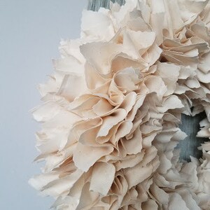 May include: A cream-colored fabric wreath with a ruffled texture. The wreath is made of many layers of fabric, creating a full and fluffy look.