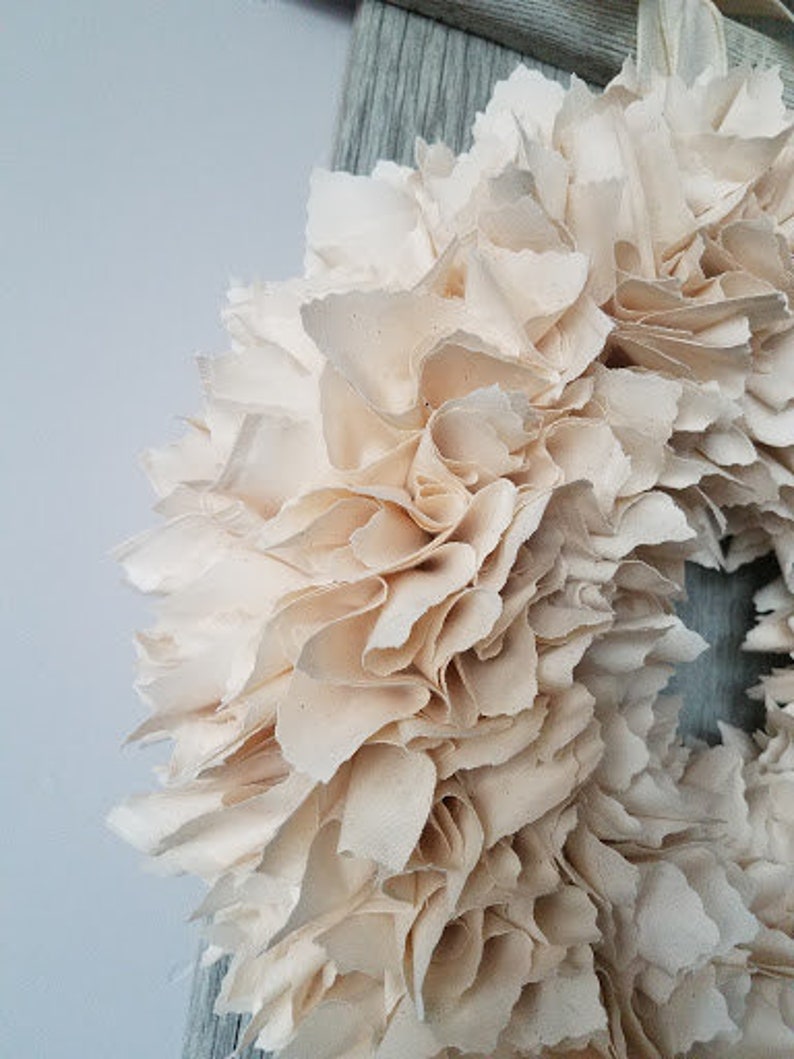May include: A cream-colored fabric wreath with a ruffled texture. The wreath is made of many layers of fabric, creating a full and fluffy look.