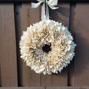 May include: A cream-colored paper flower wreath with a burlap bow hanging on a brown wooden door.