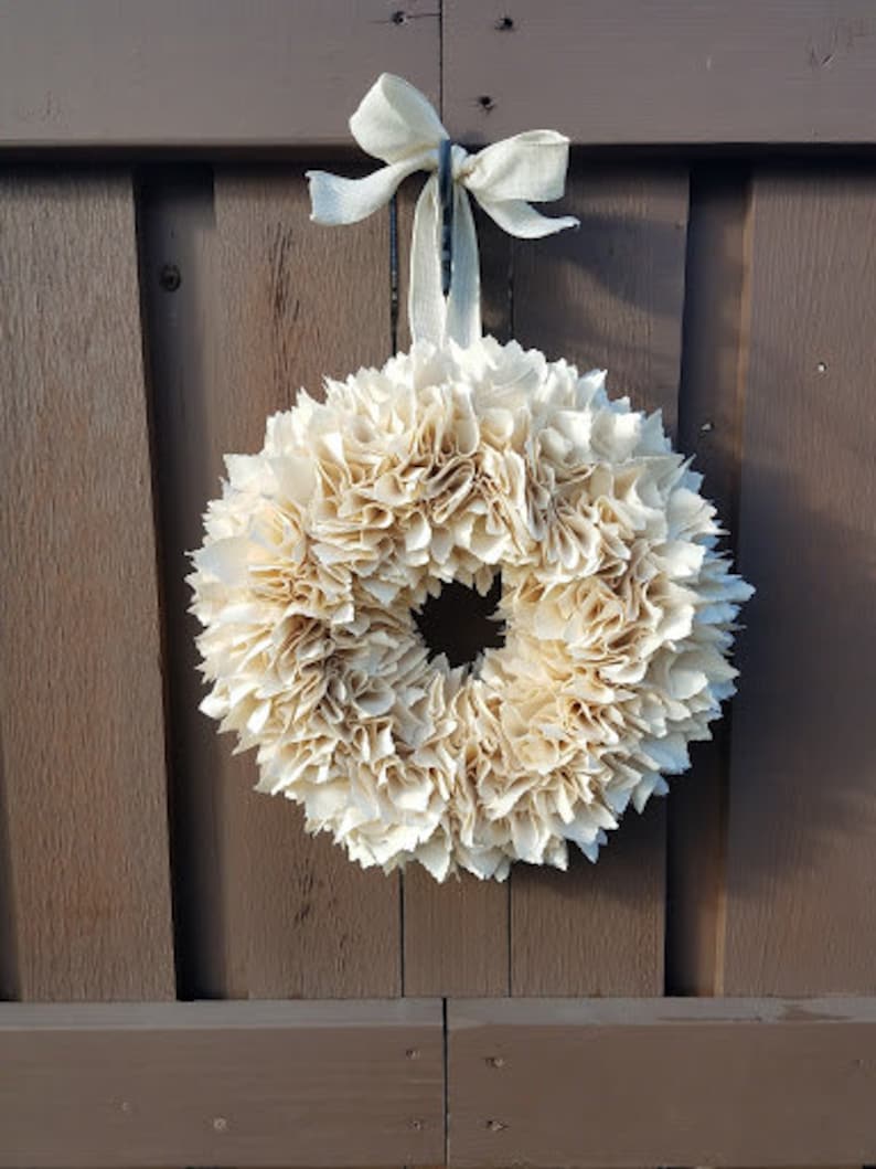 May include: A cream-colored paper flower wreath with a burlap bow hanging on a brown wooden door.