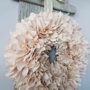 May include: A cream-colored fabric wreath with a ruffled texture. The wreath is hanging on a wooden door.