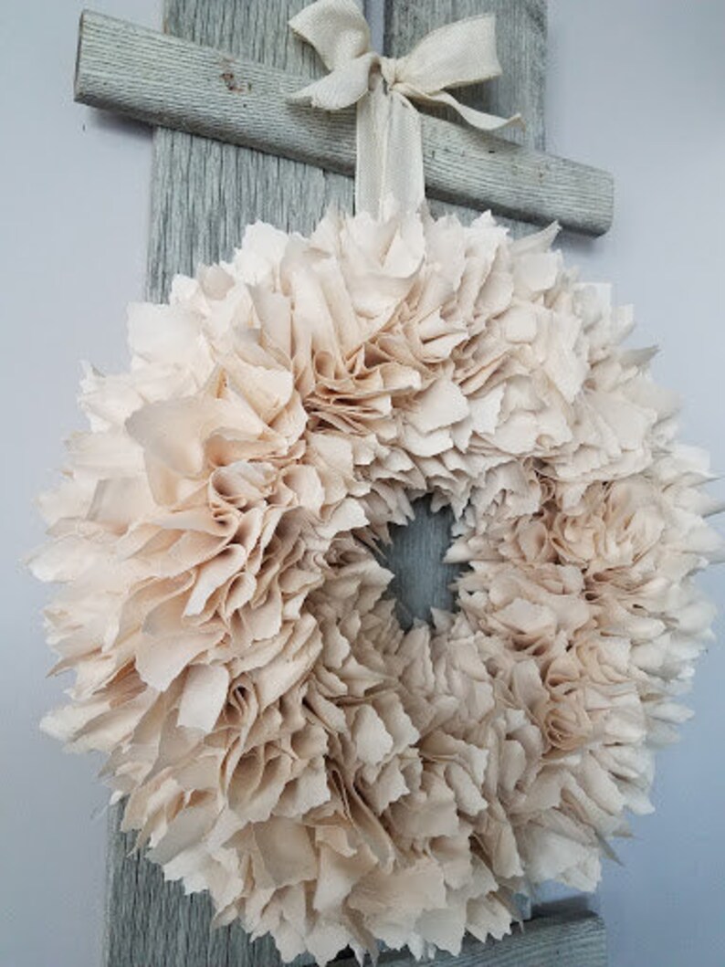 May include: A cream-colored fabric wreath with a ruffled texture. The wreath is hanging on a wooden door.