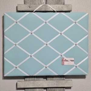 May include: A light blue fabric bulletin board with white ribbon crisscrossing the surface. The board is decorated with white pearl-like buttons.