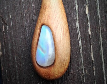 May include: A wooden teardrop pendant with a rainbow moonstone inlay. The pendant has a silver bail and is hanging against a dark wood background.