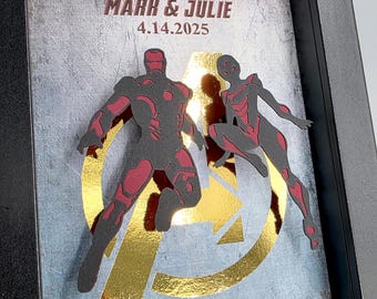 May include: A framed 3D layered art piece featuring silhouettes of two armored figures in dark gray and red over a metallic gold Avengers logo. The background is a textured gray surface with the text MARK & JULIE 4.14.2025 and I LOVE YOU 3000.