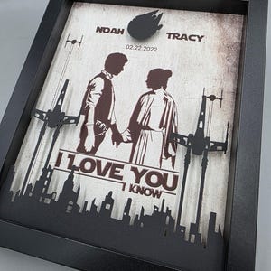 May include: A black framed shadow box featuring a layered paper cutout design. The artwork displays silhouettes of two figures holding hands, spaceships, and a city skyline. Text includes NOAH, TRACY, 02.22.2022, I LOVE YOU, and I KNOW.