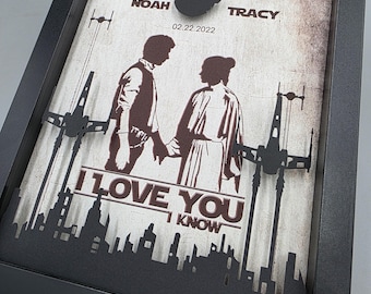 May include: A black framed shadow box featuring a layered paper cutout design. The artwork displays silhouettes of two figures holding hands, spaceships, and a city skyline. Text includes NOAH, TRACY, 02.22.2022, I LOVE YOU, and I KNOW.