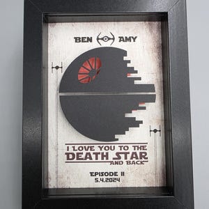 May include: A black framed shadow box featuring a layered paper cutout of the Death Star in dark gray. The background includes the text BEN, AMY, and I LOVE YOU TO THE DEATH STAR AND BACK, EPISODE II, 5.4.2024, with small TIE fighter silhouettes.