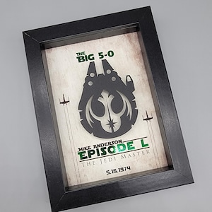 May include: A black framed shadow box containing a layered paper art piece. The design features a dark silhouette of a spaceship with a Rebel Alliance symbol, flanked by two small starfighters. Text reads: THE BIG 5-0, MIKE ANDERSON, EPISODE L, THE JEDI MASTER, 5.15.1974.