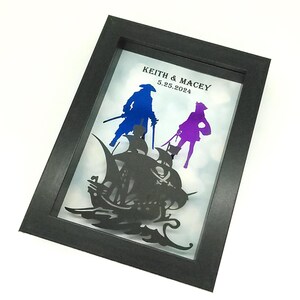 May include: Framed artwork featuring a pirate ship silhouette with two figures in blue and purple. The figures are holding swords. The text "KEITH & MACEY 5.25.2024" is displayed above the figures. The frame is black.