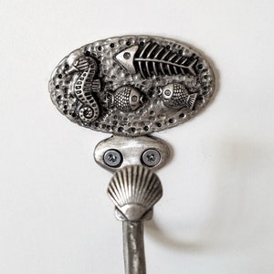 May include: Silver hook with a seashell design. The hook is attached to a decorative plate with a textured surface. The plate features a seahorse, a fish skeleton, and two fish.