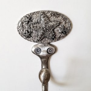 Könnte beinhalten: Silberner Haken mit einem Seestern-Design. Der Haken ist mit zwei Schrauben an einer Wand befestigt.